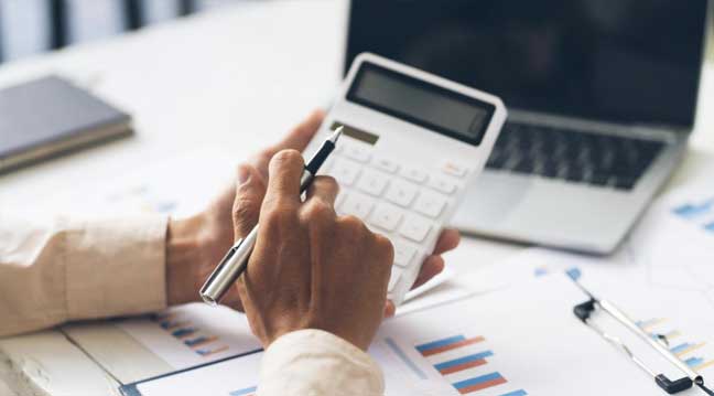 Business professional using calculator to calculate excise duty payments with financial reports and laptop for monthly compliance reporting.